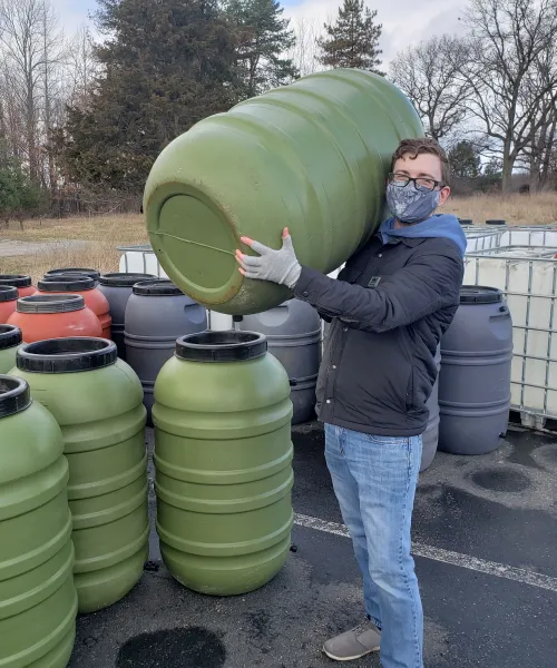 An adult wearing a mask, holding a rain barrel, next to a collection of rain barrels.