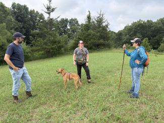 three people stand in a clearing in a nature preserve with a dog