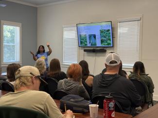 people seated in a room with their backs to the camera while watching a presentation being given on a tv screen