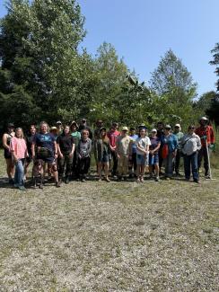 a large group of people stands in a clearing of a field with one dog and a cutting of Tree of Heaven 
