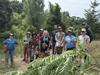 group of people stand together in a field facing the camera with a large branch of tree of heaven