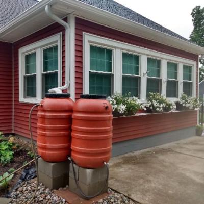 Terra cotta Rain barrels on cinder blocks in front of red house with flower garden & window boxes