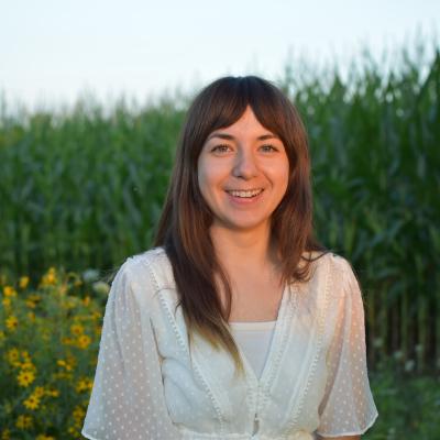 WCCD staff member Kristen Sutter standing in front of pollinator habitat and a cornfield in a white shirt.