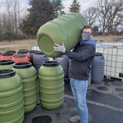 An adult wearing a mask, holding a rain barrel, next to a collection of rain barrels.